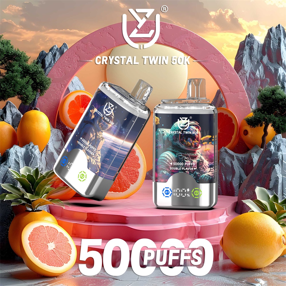 Two metallic Crystal Twin 50K disposable vape devices displayed on a pink circular pedestal surrounded by halved grapefruits, whole oranges and stylized rocky trees, with large 50000 PUFFS text and a sunset sky backdrop.