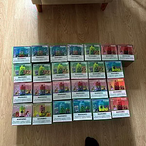 Rows of colorful Bang disposable vape pod boxes with prominent nicotine-warning labels arranged in a grid on a wooden floor under a sofa.