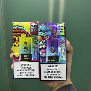 Hand holding two colorful Bang King disposable vape boxes—one yellow-green and one purple-blue with stylized gorilla artwork and brand logos, each bearing a prominent warning that the product contains nicotine and that nicotine is an addictive chemical.