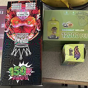Three boxed disposable vape devices on a countertop — a large black 'Bang Legend' box with red mask artwork labeled '150K Puffs', a green 'Coconut Melon 12000 Puffs' box, and a small colorful 'War' box, with bananas visible at the top edge.