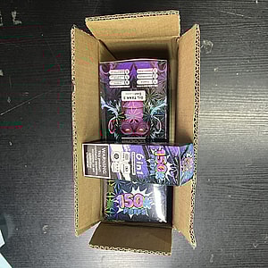 Top-down view of an open corrugated cardboard box on a dark table containing three colorful holographic disposable vape product boxes, one labeled "150k Puffs" and another "6 in 1," with warning labels visible.