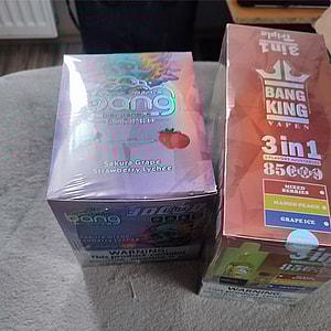 Two sealed disposable vape boxes on a gray fabric surface: a purple iridescent box labeled Sakura Grape Strawberry Lychee on the left and an orange 'Bang King 3-in-1 85 puffs' box listing Mixed Berries, Mango Peach, and Grape Ice on the right.