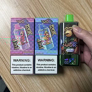 Two colorful boxed vape packages—one purple and one blue—each showing product artwork and a prominent nicotine warning label at the bottom, placed on a wood surface next to a green handheld vape device with a cartoon gorilla graphic and a green cap being held by a hand.