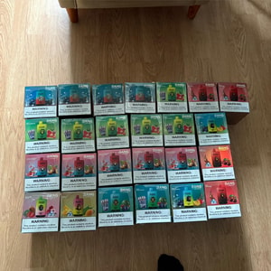 Rows of colorful Bang disposable vape pod boxes with prominent nicotine-warning labels arranged in a grid on a wooden floor under a sofa.