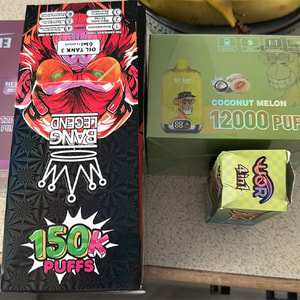 Three boxed disposable vape devices on a countertop — a large black 'Bang Legend' box with red mask artwork labeled '150K Puffs', a green 'Coconut Melon 12000 Puffs' box, and a small colorful 'War' box, with bananas visible at the top edge.