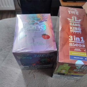 Two sealed disposable vape boxes on a gray fabric surface: a purple iridescent box labeled Sakura Grape Strawberry Lychee on the left and an orange 'Bang King 3-in-1 85 puffs' box listing Mixed Berries, Mango Peach, and Grape Ice on the right.