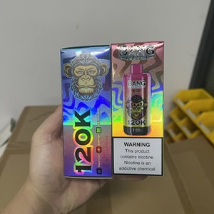 Hand holding two holographic vape product boxes — one with large iridescent '120K' text and the other showing a pink 'Bang Legend' bottle image and a nicotine warning label, set against a neutral indoor background with cardboard and yellow plastic bins.